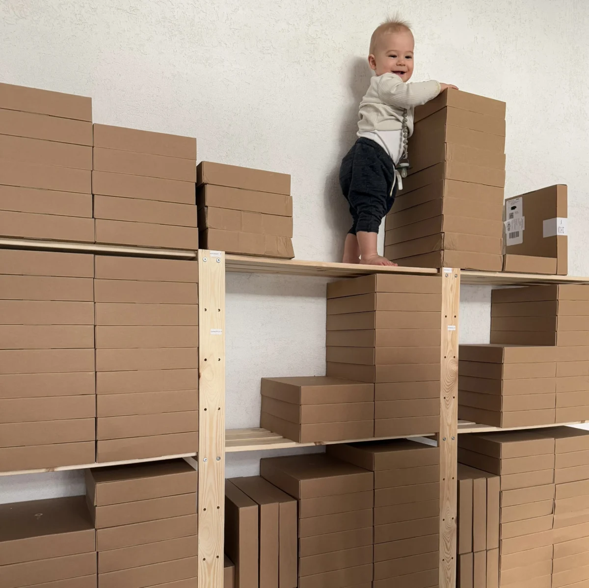 Unsere Vision 1 Ein freudiges Kleinkind steht bei Mriella neben ordentlich gestapelten Versandkartons der Babyalben – passend zum Slogan "Ein Leben. Box für Box."